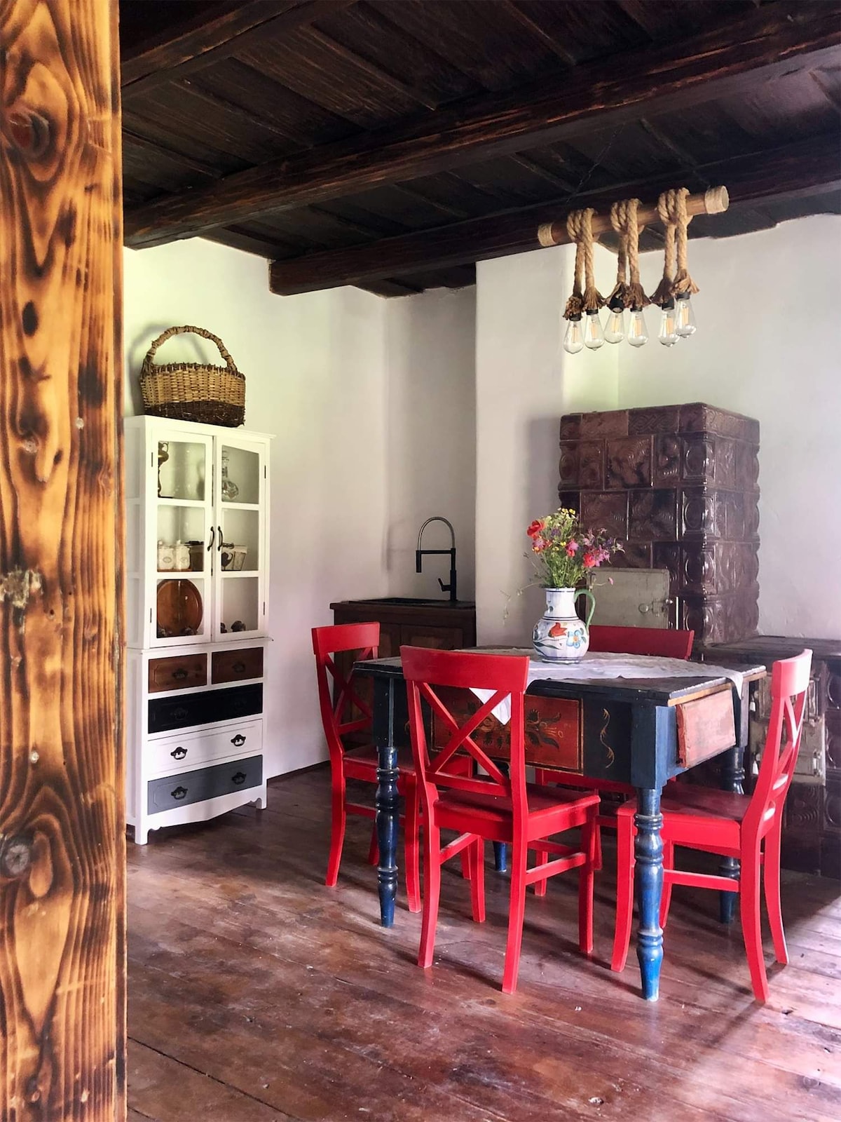 A rustic dining area features a wooden table surrounded by red chairs. An antique cupboard stands against the wall, displaying various items. Above, a unique light fixture made of ropes and bulbs adds character to the space, which is illuminated by natural light.