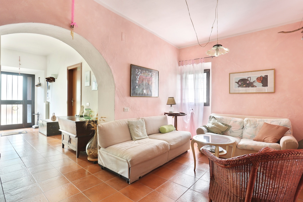 A living area features light pink walls and terracotta tile flooring. A spacious sofa and a wicker chair are arranged around a small table. Natural light filters through sheer curtains, illuminating the space. An arched doorway leads to another room, adding architectural interest.