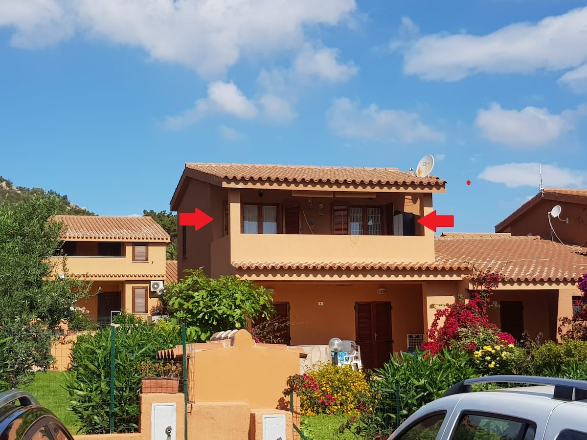 The image captures the exterior of a two-story villa, characterized by a terracotta roof and light-colored walls. Windows on the upper floor allow natural light indoors, while vibrant greenery and flowers are present in the surrounding garden area. Nearby parking spaces are visible.