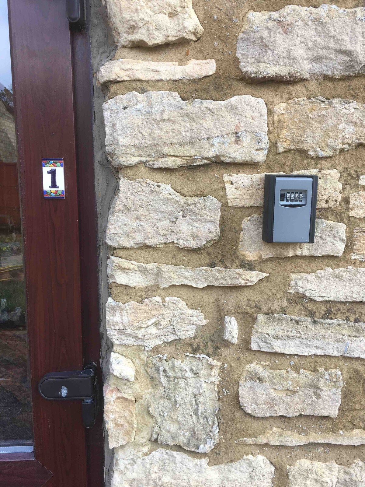 The entrance area features a stone wall composition with natural textures and a secure keyless entry system. A wooden door with a dark finish provides access, and a number '1' is displayed on a colorful plaque, indicating the unit's designation.