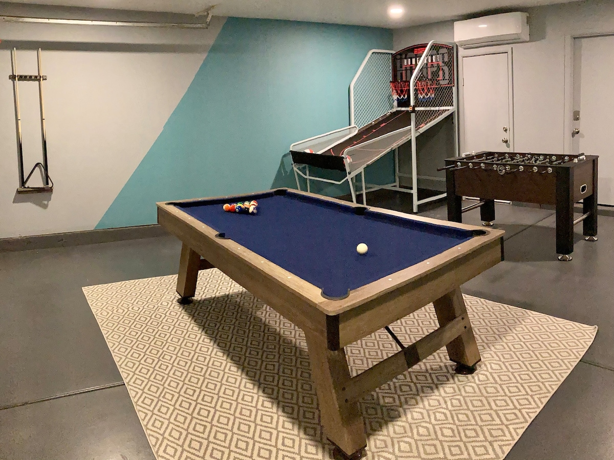 A game room is visible, featuring a billiards table with a blue felt surface and a set of colorful pool balls. A foosball table is situated nearby, and a dual hoop basketball game stands against the wall. The room is painted in a calming blue hue, complemented by a geometric patterned rug.