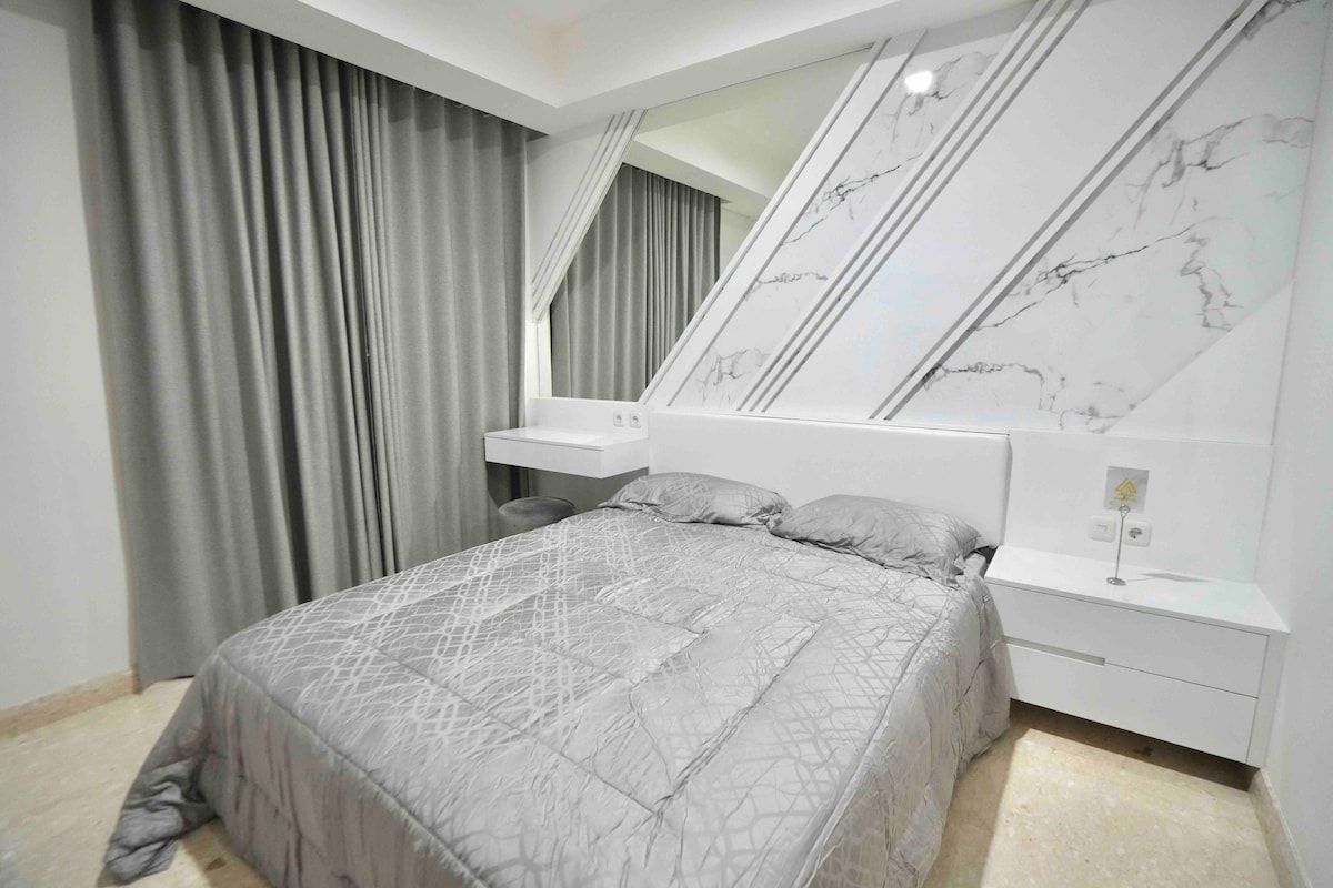 A spacious sleeping area features a large bed dressed in a soft gray quilt. A sleek white bedside table is positioned next to the bed, and the room is enhanced by floor-to-ceiling curtains in a complementary shade, with a geometric marble accent wall adding visual interest.