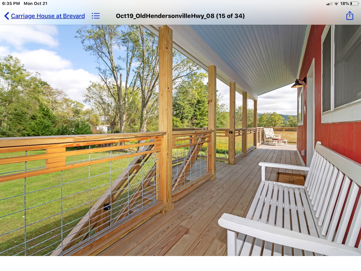 A covered porch features wooden decking and a railing with horizontal metal accents. Seating is provided by a white bench and two lounge chairs facing the expansive green lawn and surrounding trees, creating a serene outdoor space.