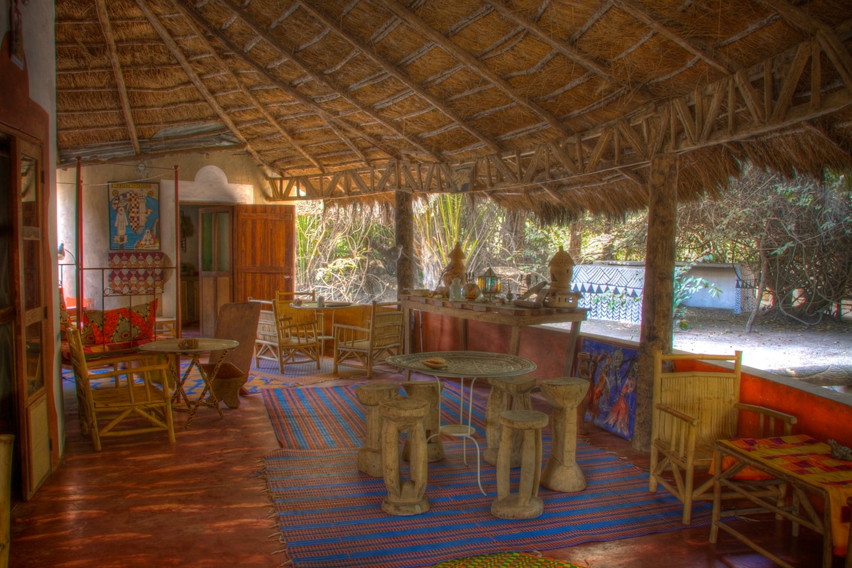 The spacious outdoor living area features a thatched roof supported by wooden beams. Wooden tables and chairs are arranged on a vibrant woven rug. Natural light filters in, illuminating the surrounding greenery, creating a welcoming space for relaxation and social gatherings.