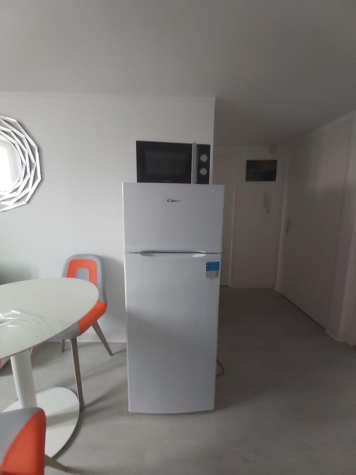 A modern, upright refrigerator stands against a white wall, reflecting its sleek design. Above the refrigerator, a compact microwave is mounted. A circular dining table with orange and white chairs is positioned nearby, contributing to the clean and functional atmosphere of the space.