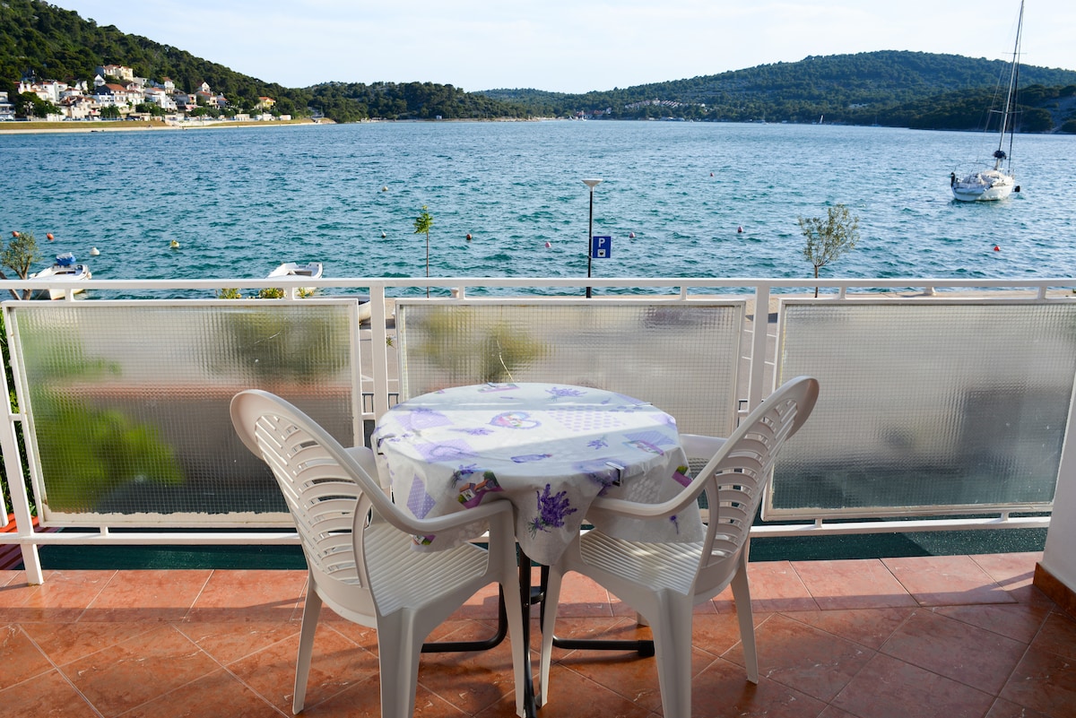 A spacious balcony presents a round table set with a floral tablecloth, accompanied by two lightweight chairs. The view showcases a serene body of water bordered by lush hills and distant homes, enhancing the outdoor space's natural tranquility.