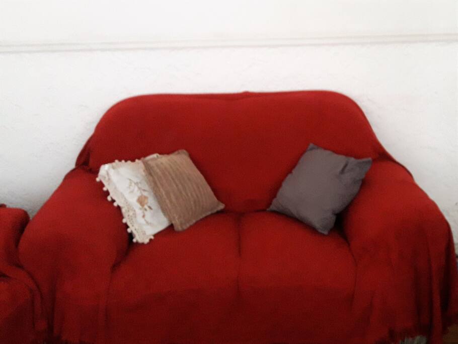 Large living room with two large sofas and space to put another mattress on the floor. It has a wooden furniture, two large windows and a round glass table with 4 chairs