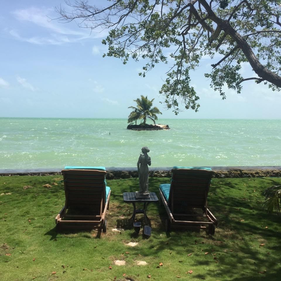 Two wooden lounge chairs are positioned on a grassy area, facing the ocean. A small island with a palm tree is visible in the distance, framed by gentle turquoise waters. A statue stands on a side table between the chairs, enhancing the serene setting.