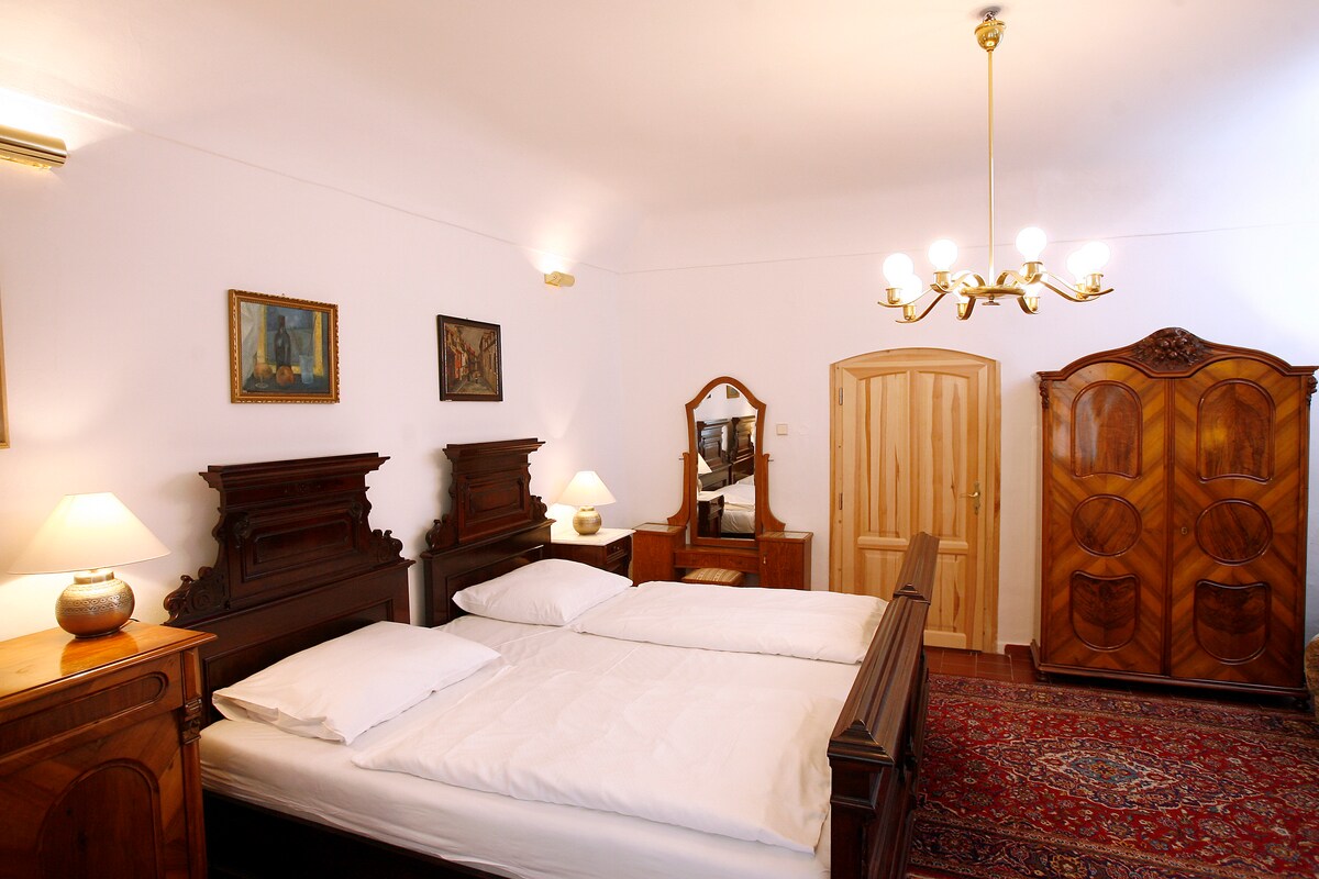 The main bedroom is furnished with a spacious bed and decorative wooden furniture, including a tall wardrobe and a mirror. Soft lighting is provided by bedside lamps and a chandelier, illuminating the room's elegant decor and creating a warm ambiance.