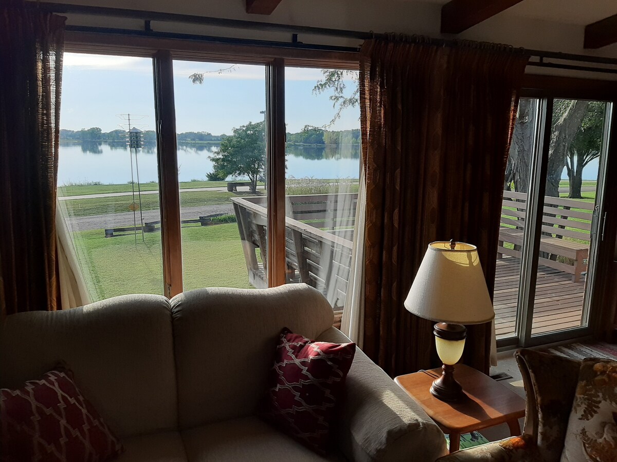 Large windows frame a serene view of Augusta Lake, allowing natural light to illuminate the living space. A comfortable sofa with decorative pillows faces the windows, and a lamp on a side table provides additional lighting. The outdoor deck is visible through the glass doors.