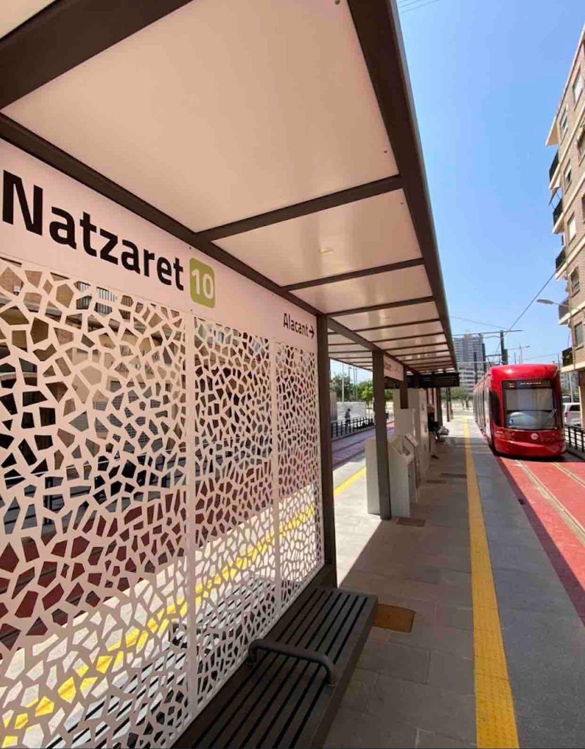 A modern tram station features a covered waiting area with a patterned screen providing shade. A tram in red awaits on the tracks, with visible signage indicating the Natzaret line. Clear blue skies can be seen above, enhancing the bright setting.