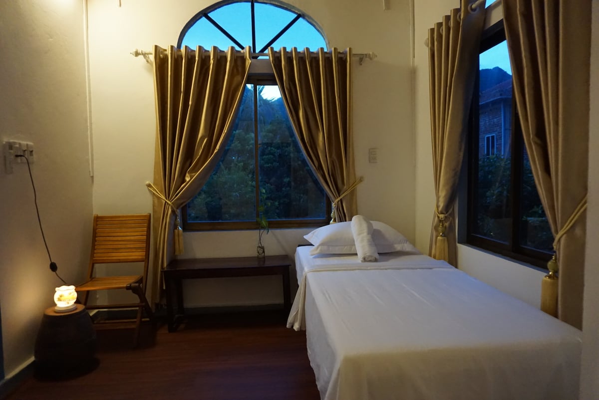 A bright and airy guest room features a large window with scenic views, dressed with soft drapes. One double bed and one single bed are arranged with crisp linens. A wooden chair and table complement the space, while a small lamp adds a gentle glow.