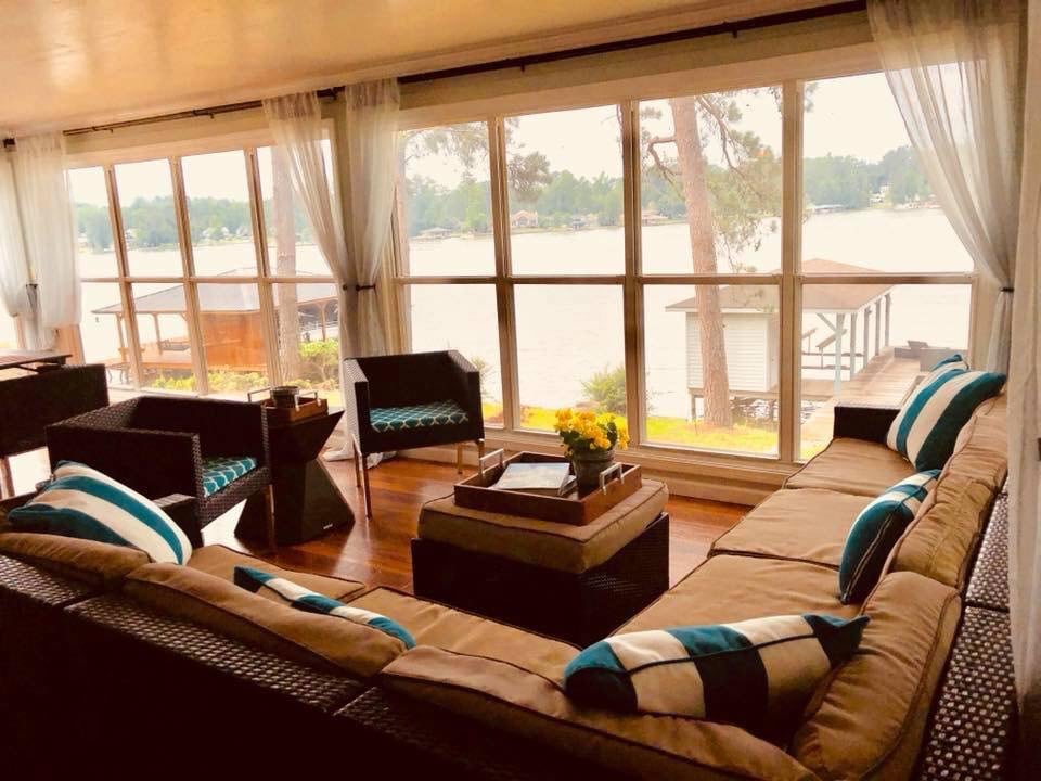 A spacious sunroom features large windows offering a panoramic view of the lake. The seating area is arranged with a sectional sofa adorned with blue and white cushions, complemented by decorative tables. Soft curtains frame the windows, enhancing the bright, airy space.