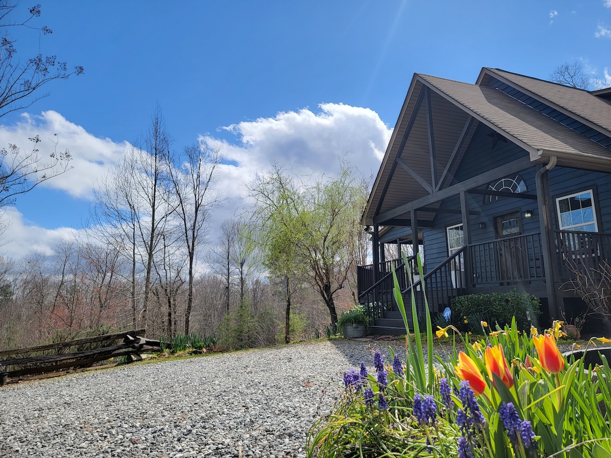 The modern cottage is nestled among trees under a clear blue sky, with fluffy white clouds visible. A gravel pathway leads to the entrance, framed by vibrant flowers, including tulips and grape hyacinths, adding color to the serene surroundings.