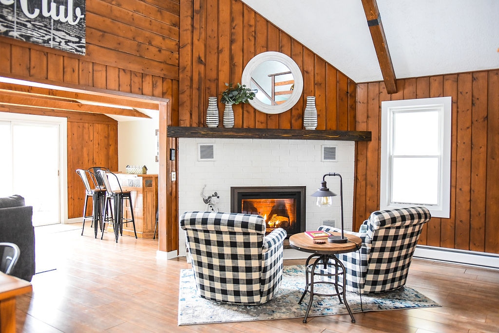 Two comfortable checkered armchairs are positioned around a small round table in front of a white-brick fireplace. Warm wood paneling covers the walls, complemented by light from windows on either side. A rustic mirror is mounted above the fireplace, enhancing the cozy ambiance.