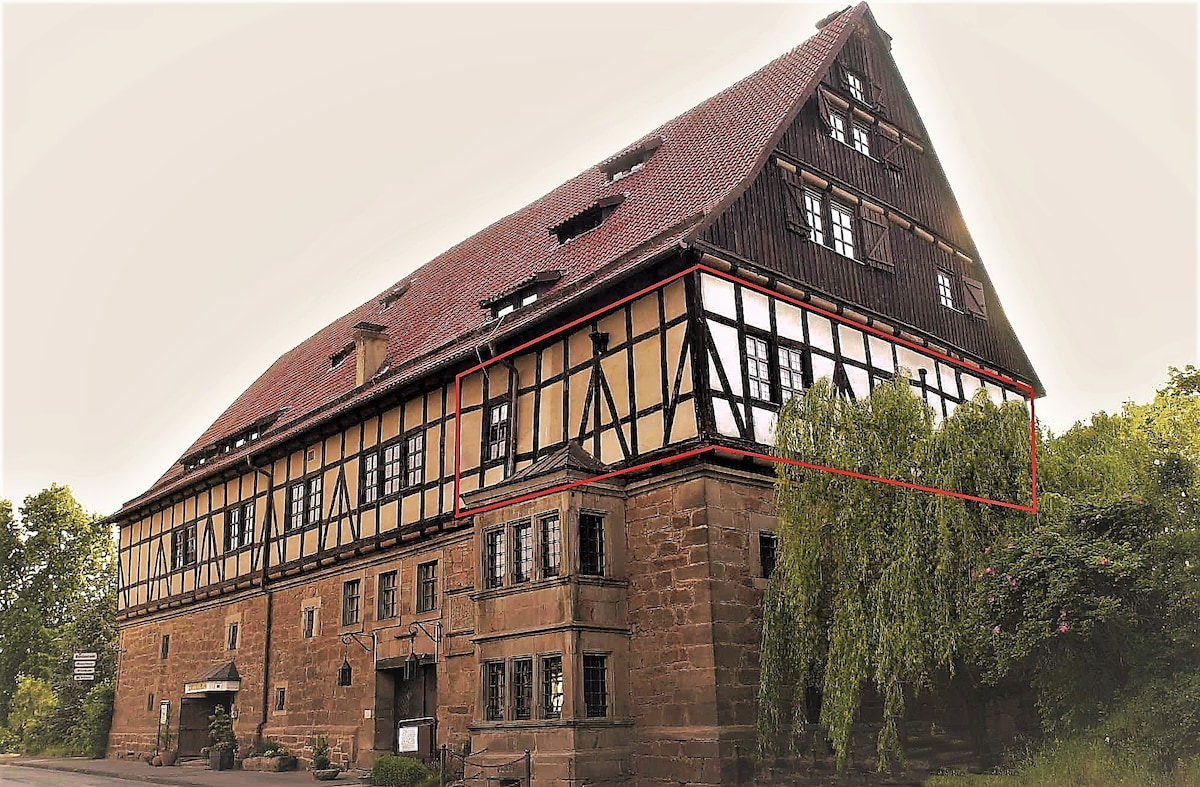 The historic building features a striking mix of timber-framed and stone architecture, showcasing a blend of craftsmanship from its long history. Large windows allow natural light to illuminate the space, while lush greenery surrounds the property, enhancing its inviting exterior.