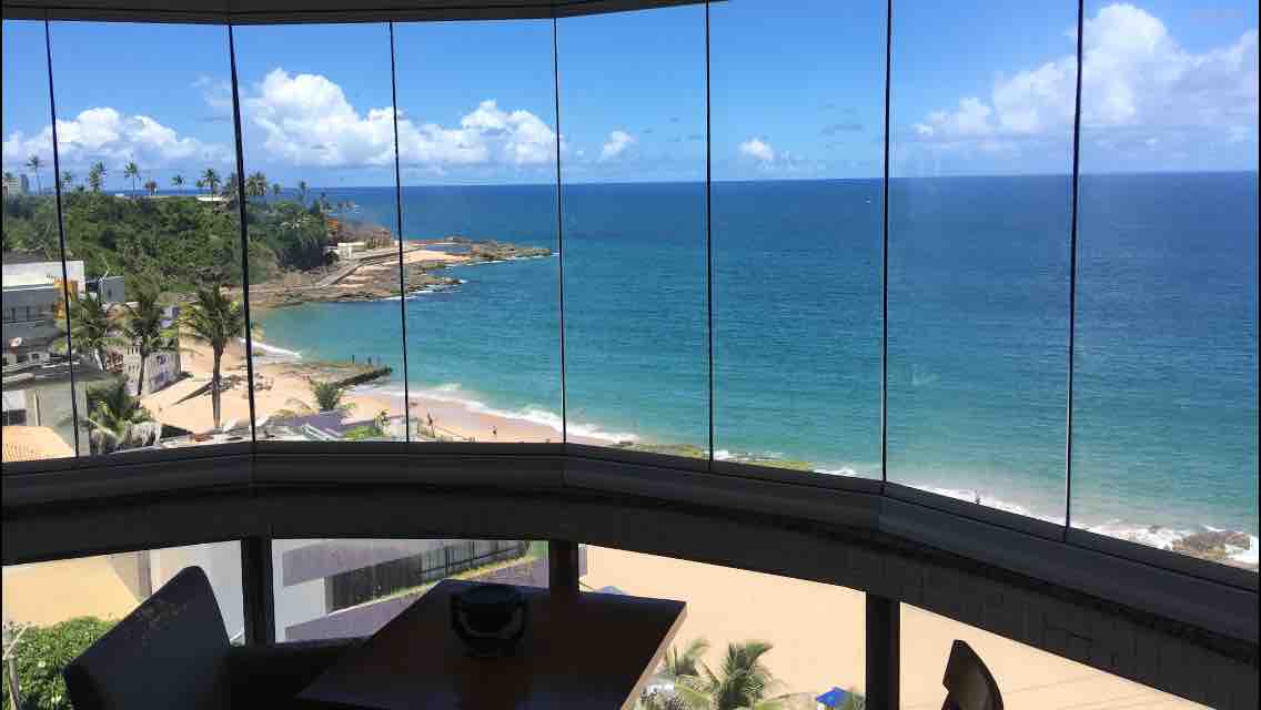 A panoramic window offers a stunning view of the turquoise ocean meeting the sandy beach. Palm trees frame the scene, and the shoreline extends along a sunny coastline under a bright blue sky with scattered clouds.