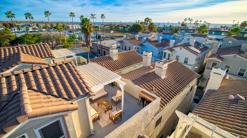 An aerial view captures the rooftops of surrounding homes, highlighting the cozy rooftop deck with comfortable seating. The ocean and palm trees are visible in the distance, framing a serene coastal atmosphere.