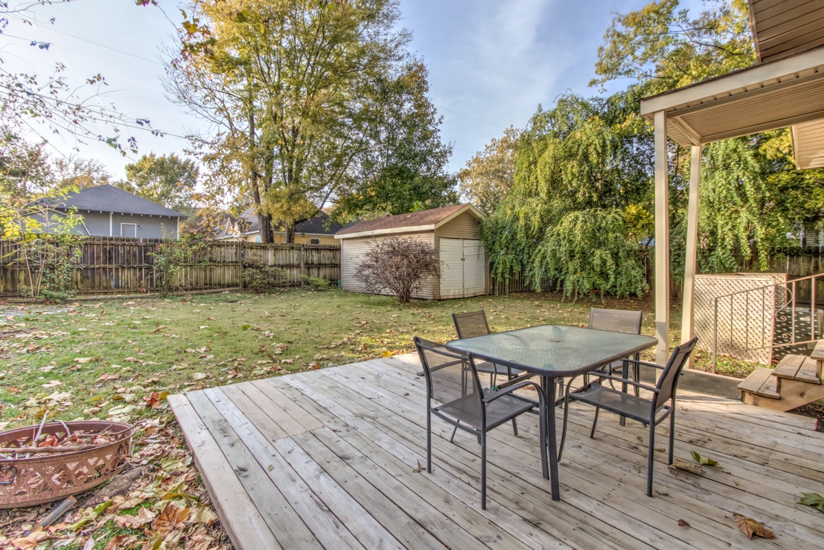 A spacious wooden deck extends into a large backyard, featuring a glass-top table surrounded by six black chairs. Lush greenery surrounds the area, with several trees providing shade. A small shed is visible in the background, and fallen leaves cover the ground.