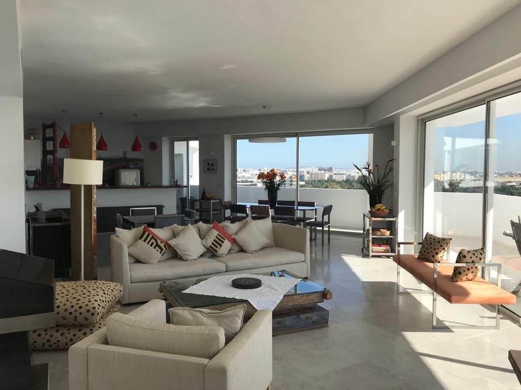 The spacious living area features large floor-to-ceiling windows that allow abundant natural light. A modern sofa with decorative cushions is positioned in the center, accompanied by a stylish coffee table. In the background, the dining area and kitchen are visible, enhancing the open concept design.