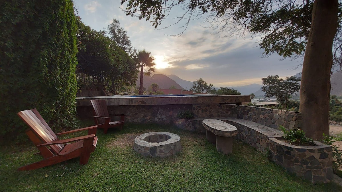 An outdoor seating area features wooden lounge chairs and stone benches surrounding a circular fire pit. Lush greenery and trees provide shade, while a scenic view of the mountains is visible in the background, enhancing the tranquil atmosphere.