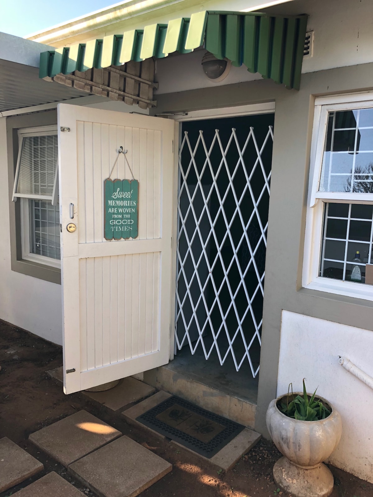 The entrance features a white door with a decorative sign that welcomes guests. A sliding security gate is positioned behind the door, ensuring safety. Light-colored walls and a green awning add to the exterior charm. A small potted plant is visible on the ground.