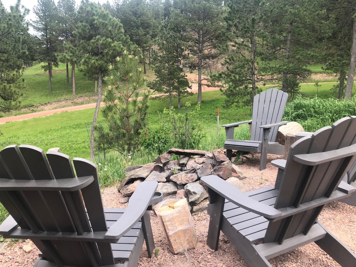 Two gray Adirondack chairs are positioned around a stone fire pit, surrounded by a mixture of gravel and grass. Tall pine trees line the background, offering a serene view of the natural landscape. A dirt path leads further into the greenery.