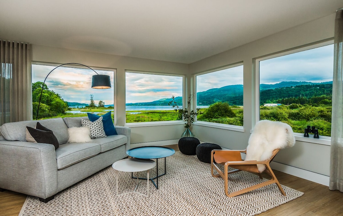 The living area showcases large panoramic windows offering views of the estuary. A comfortable sofa is accented with decorative pillows, while a cozy chair and round coffee tables are positioned on a textured area rug. Natural light enhances the serene atmosphere.
