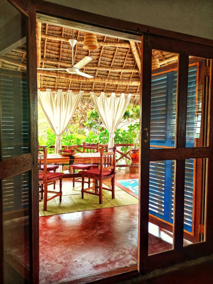 An inviting dining area is visible through open wooden doors. Natural light filters in through sheer curtains, illuminating the wooden dining table surrounded by chairs. The thatched roof adds a tropical touch, while greenery is seen through the openings, enhancing the connection to the outdoors.