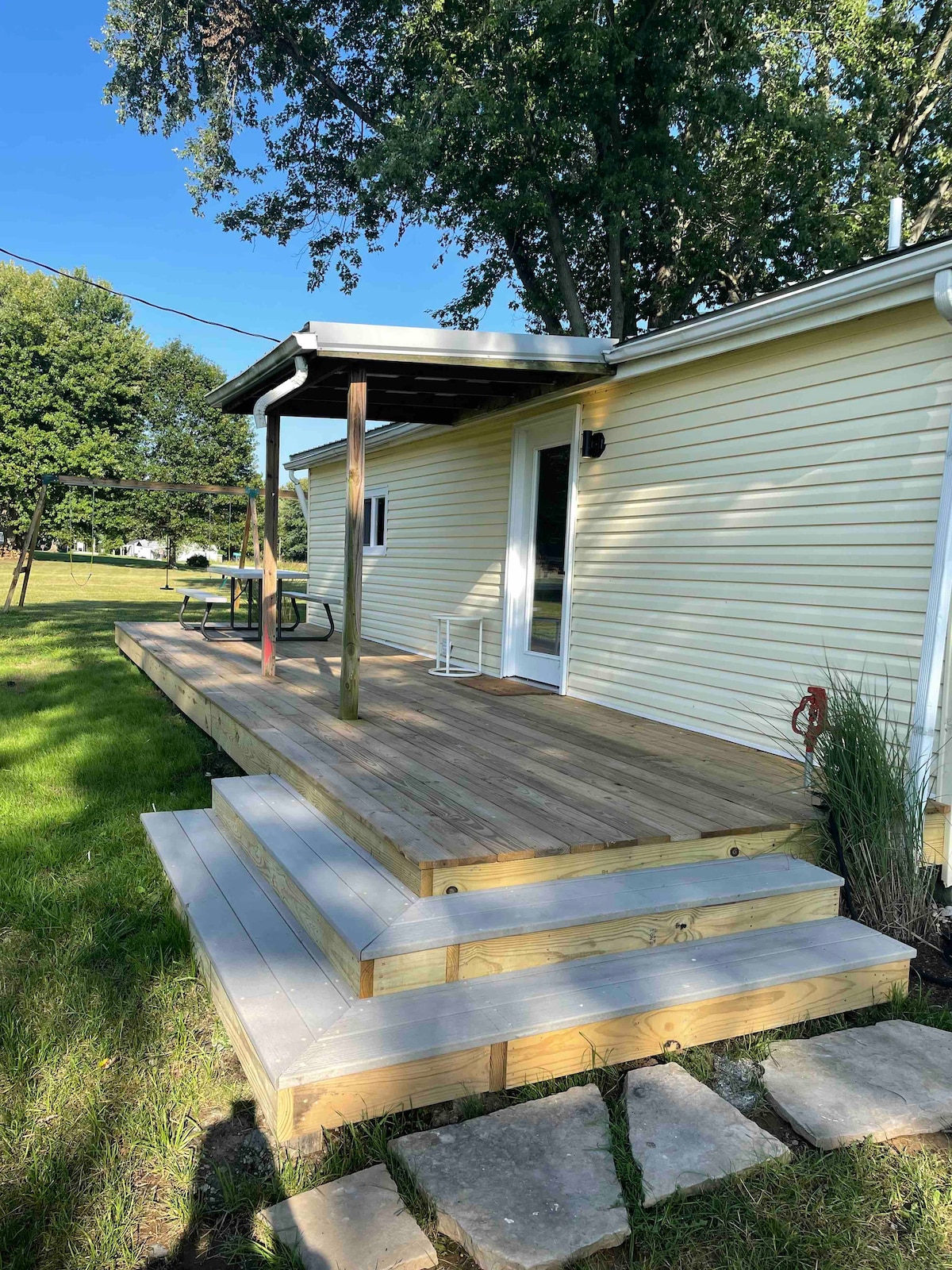 A spacious outdoor deck features wooden flooring and steps leading down to the grassy yard. A covered area provides shade, and a white rocking chair is visible. Lush greenery surrounds the home, creating an inviting space for relaxation.