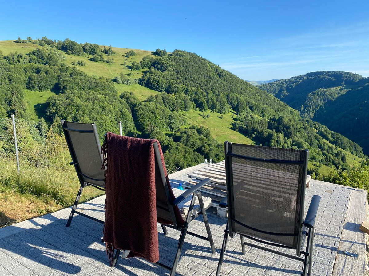 Two foldable chairs are positioned on a wooden deck, facing expansive green hills under a clear blue sky. A blanket is draped over one chair, providing a cozy touch to the serene outdoor setting.