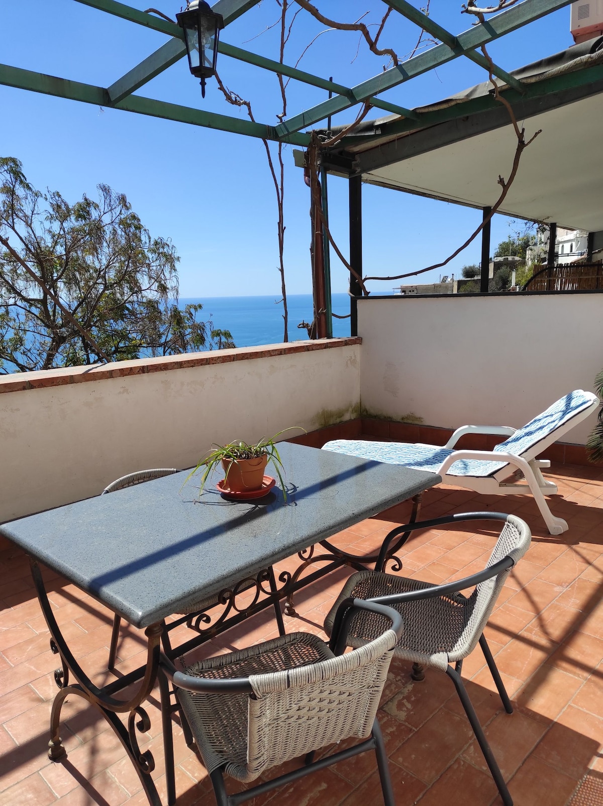 An outdoor terrace is presented, featuring a stone table surrounded by four chairs. A lounge chair is positioned beside the table. Views of the blue sea are seen in the background, while a plant pot sits atop the table, and vines are draped above the area.