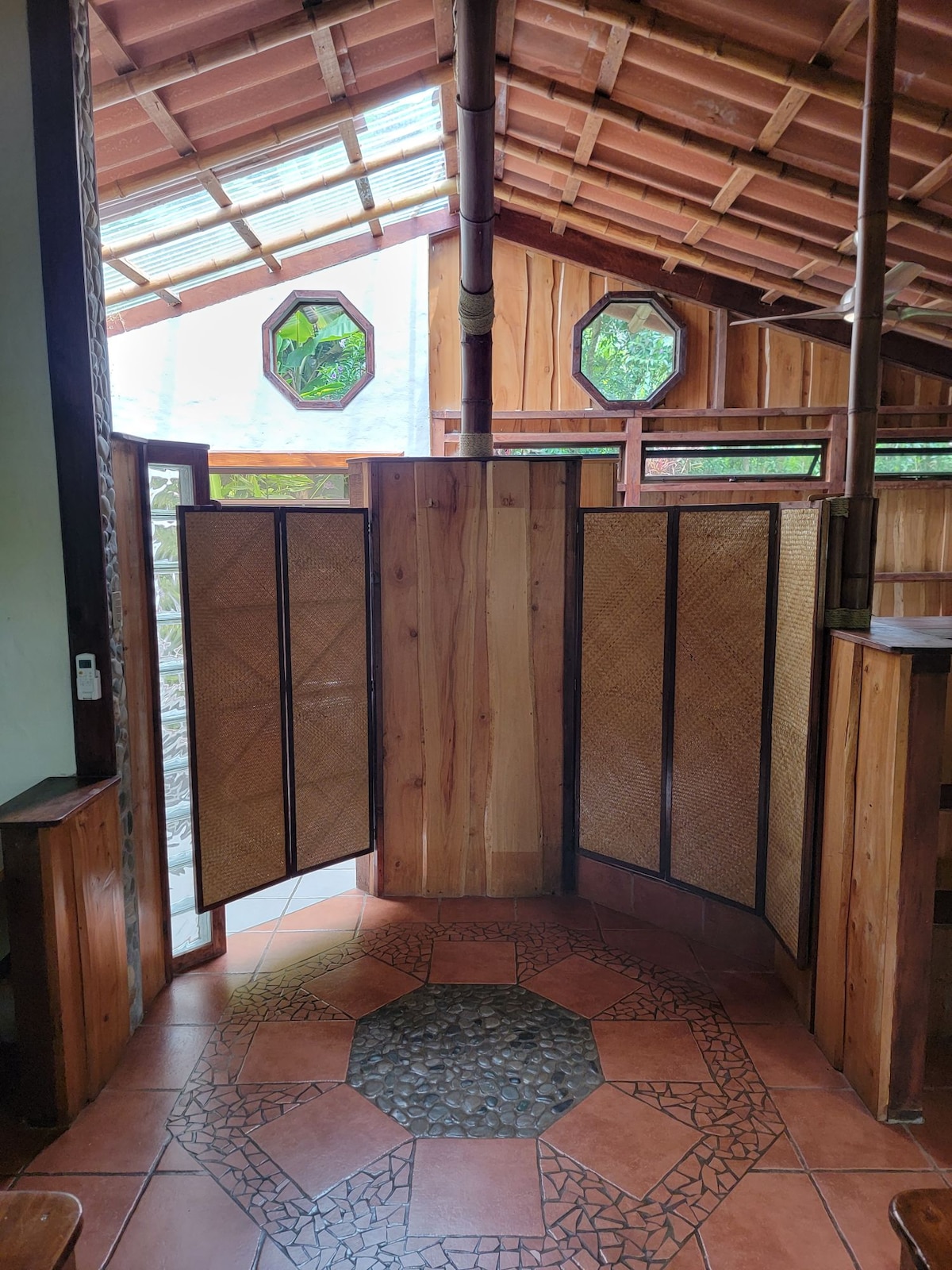 A wooden privacy screen is featured in the center of the room, made up of several folding panels. Surrounding it is an earthy tile floor with a circular stone inlay. Natural light filters through the octagonal windows above, illuminating the warm wooden structure of the space.