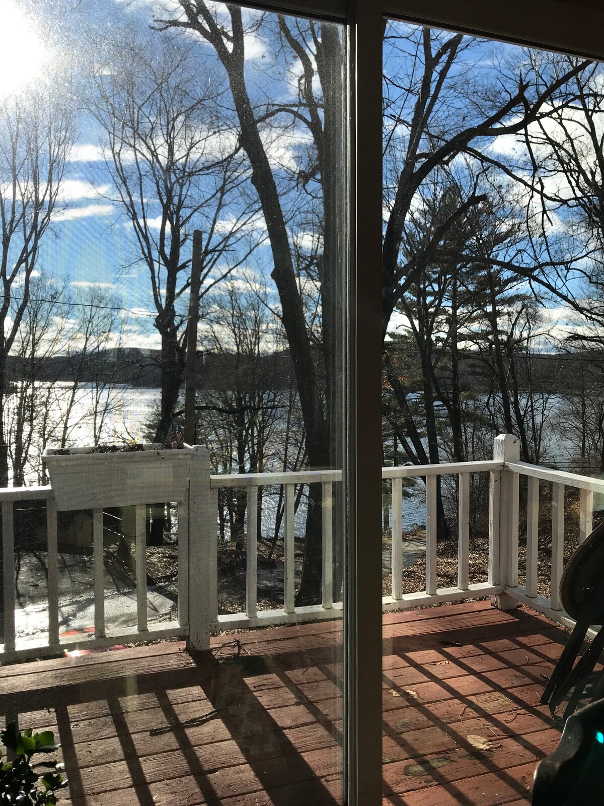 A serene view of the lake is framed by large glass doors, showcasing a deck with a wooden railing. Natural light illuminates the space, highlighting nearby trees and the shimmering water. The scene captures a peaceful moment in nature, inviting relaxation.