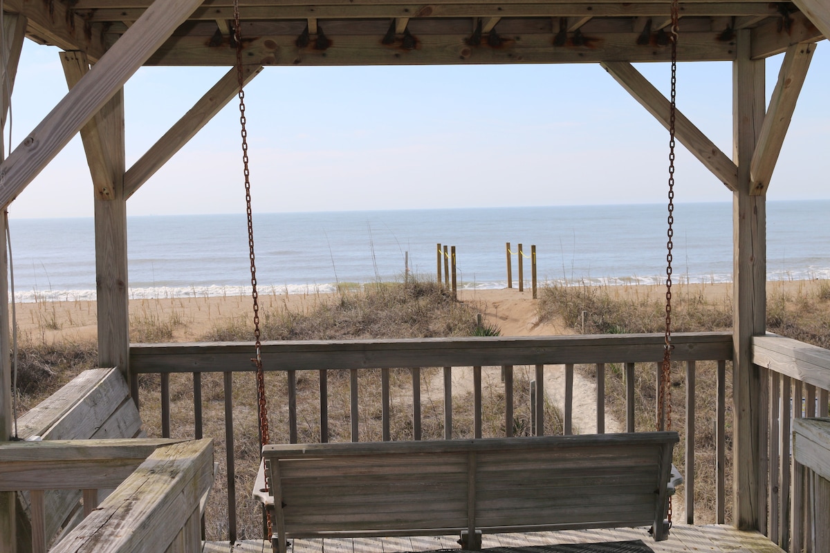 A view from a wooden gazebo features a porch swing overlooking a sandy beach and the ocean. The tranquil scene includes gentle waves lapping against the shoreline, with wooden posts visible in the distance along the beach, framed by tall grasses.