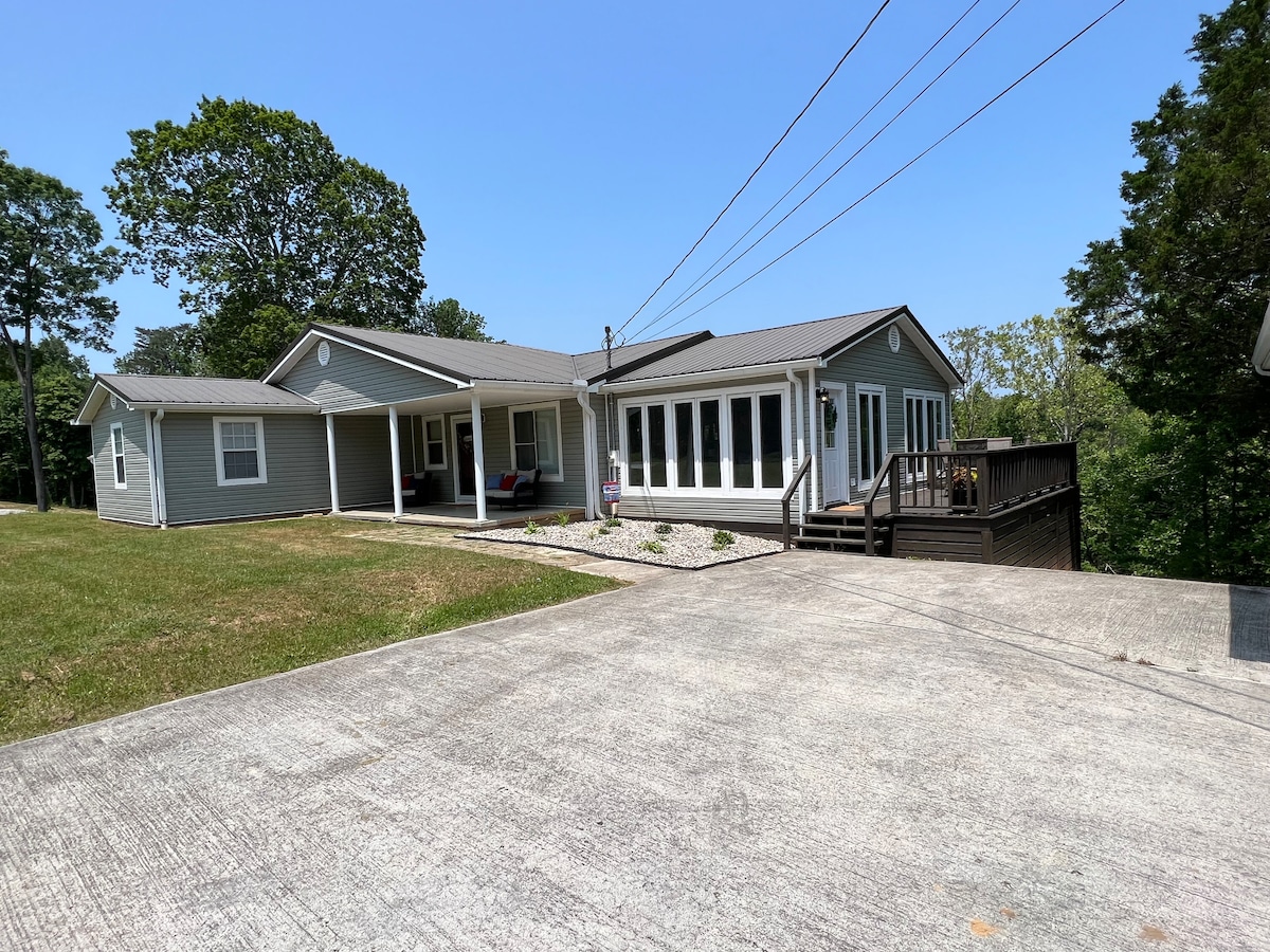 The exterior of the house showcases a spacious driveway leading up to a single-story structure. Large glass windows form a sunroom, complementing the grey siding and inviting landscape, which includes well-maintained grass and trees in the background. A wooden deck is visible in the distance.