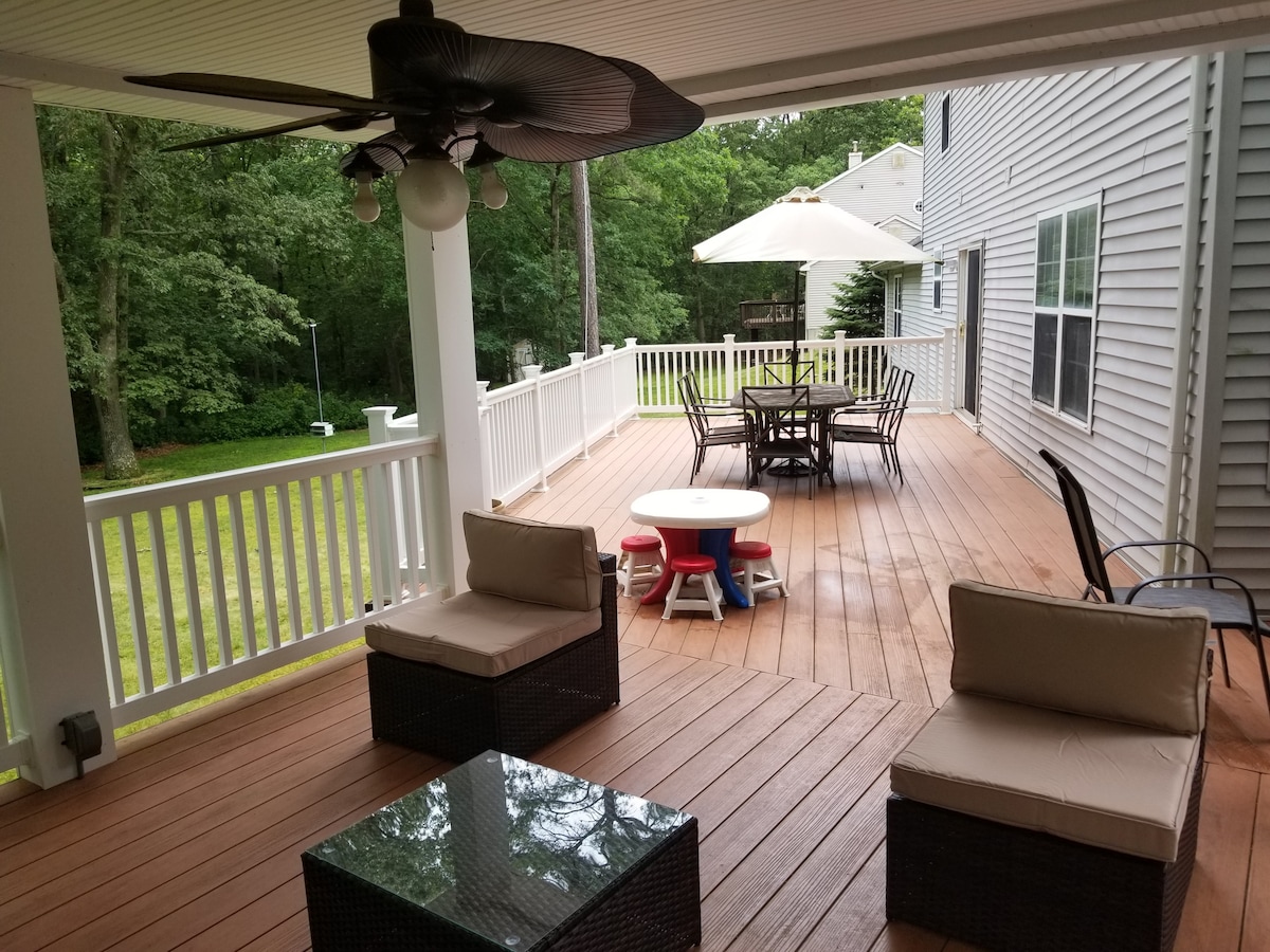 A spacious outdoor deck is captured, featuring a dining table surrounded by several chairs under a large umbrella. Two cozy seating arrangements are situated nearby, complemented by a coffee table. Lush greenery is visible in the background, creating a serene atmosphere.
