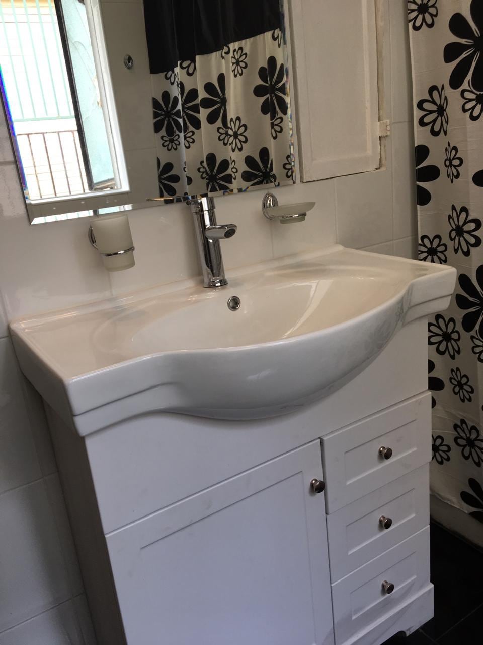 A white vanity with a curved sink is positioned against a tiled wall. A mirror is mounted above the sink, and two wall-mounted light fixtures flank either side. Floral-patterned shower curtains with black and white designs add a decorative touch.