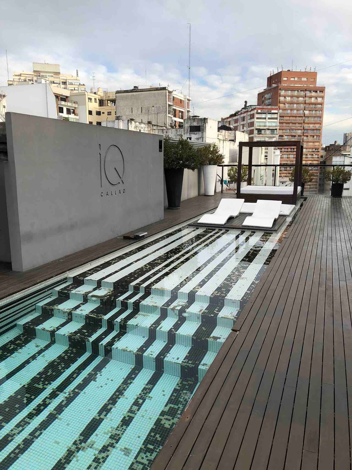 A rooftop pool is featured with a mosaic tile design, surrounded by wooden decking. Two white lounge chairs are positioned beside the pool, offering a space for relaxation. The city skyline is visible in the background, showcasing a blend of modern buildings.
