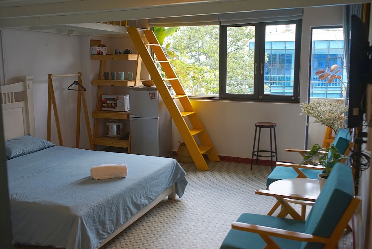 A spacious room is highlighted by a comfortable bed with a light blue coverlet. Adjacent, a ladder leads to a lofted area. Natural light fills the space through large windows, while two teal chairs and a stool provide seating. A small fridge and shelves are visible.