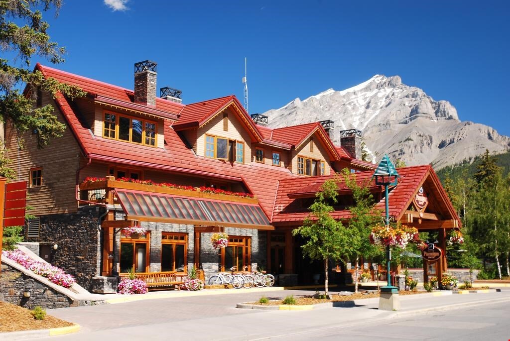 A charming mountain inn features a warm wooden exterior with red rooftops, surrounded by colorful flower boxes. In the background, a majestic snow-capped mountain rises against a clear blue sky, adding to the inviting atmosphere of the location.