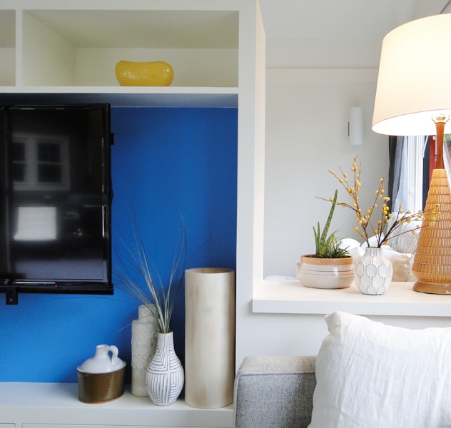 A modern living area showcases a sleek television placed against a bright blue accent wall. Various decorative elements, including potted plants and vases, add artistic flair. A warm lamp provides soft lighting, enhancing the overall comfort of the space.