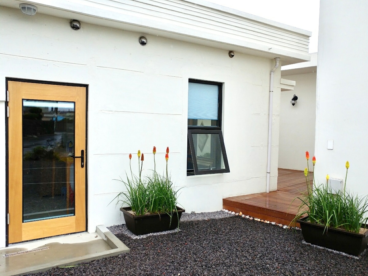 The entrance features a modern wooden door framed with decorative plants. Two planters with tall green grass and colorful flowering stems are positioned symmetrically at the front. A wooden deck is visible beside the entrance, complementing the contemporary architecture of the house.