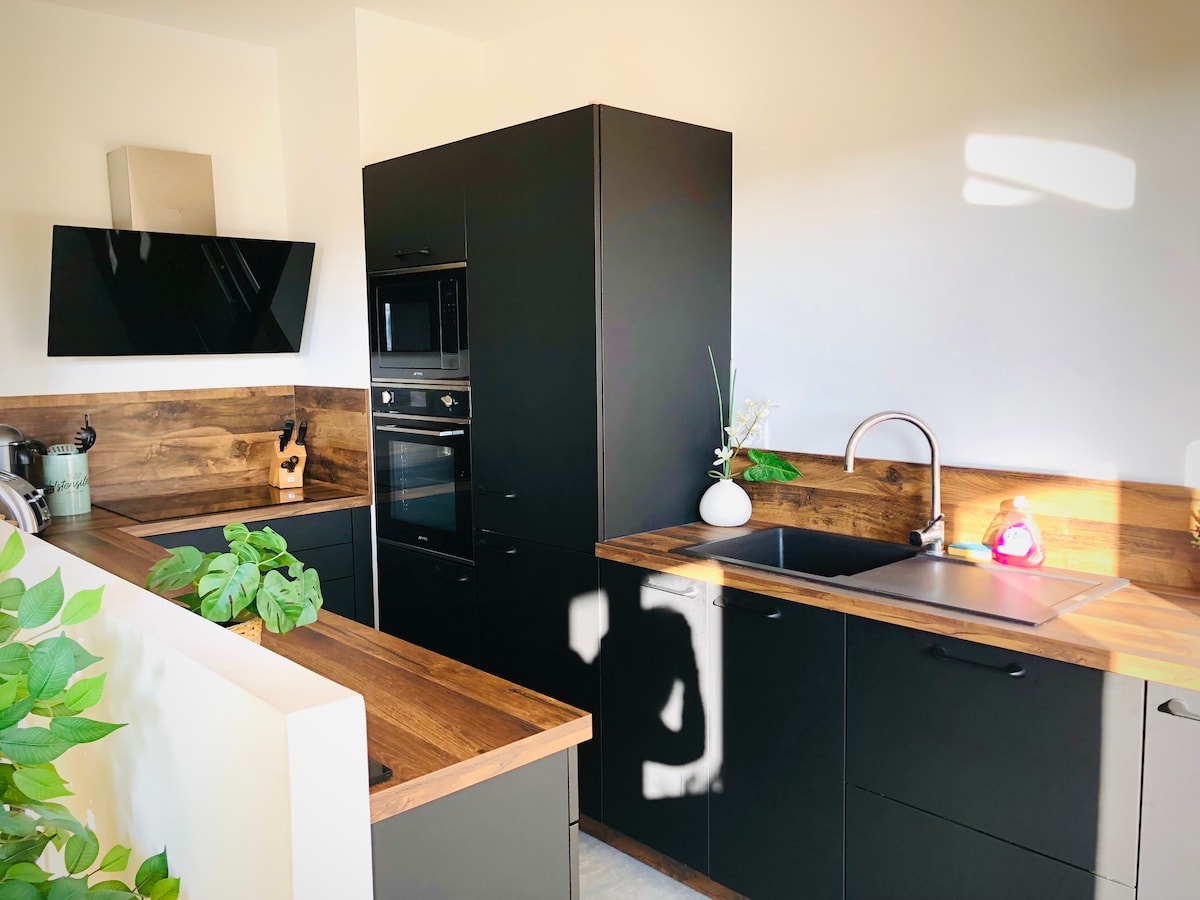 A modern kitchen is featured with sleek black cabinetry and wooden accents. Built-in appliances, including an oven and microwave, are integrated seamlessly. A sink with a minimalist faucet is present, alongside light-colored countertops. Natural light filters in, enhancing the clean and functional design.