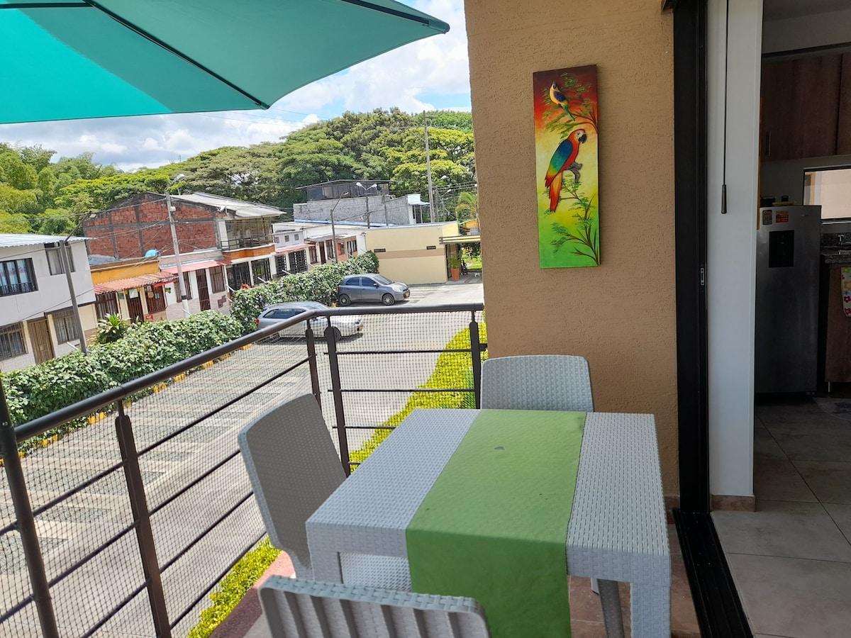 A balcony area is presented with a small dining table and four chairs. A green table runner adds a splash of color. A vibrant painting of tropical birds decorates the wall. An umbrella provides shade, while views of the street and greenery are visible beyond.