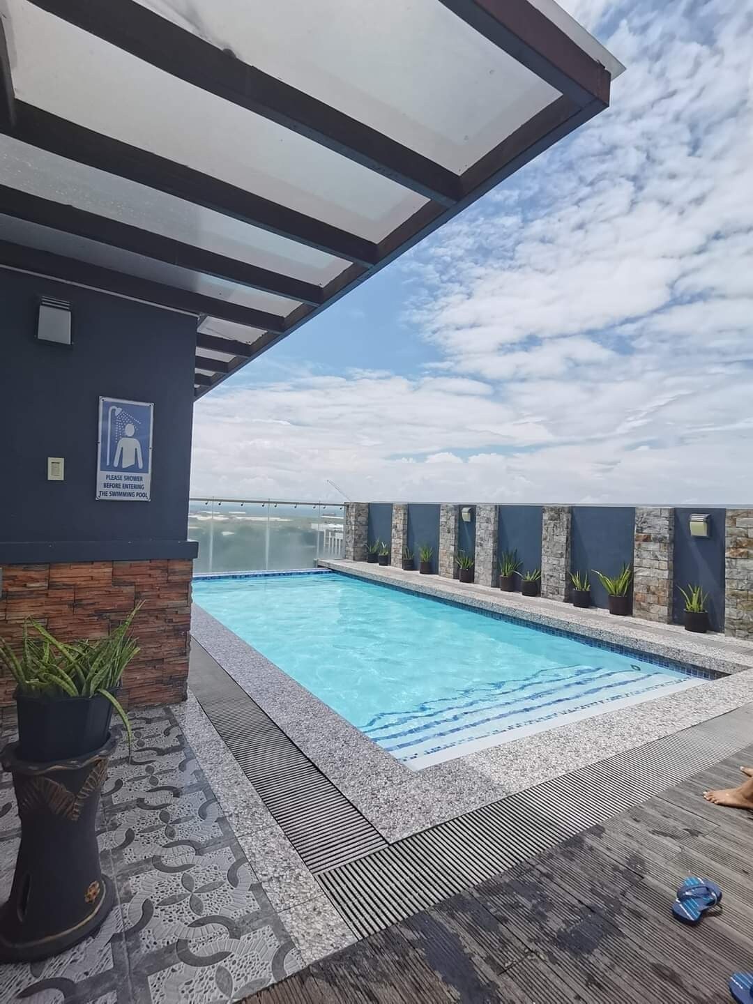 A rooftop pool is shown, surrounded by a stone wall and planters with greenery. The clear water reflects the sky above, with scattered clouds visible. Sunlight brightens the area, enhancing the calm atmosphere of the space.