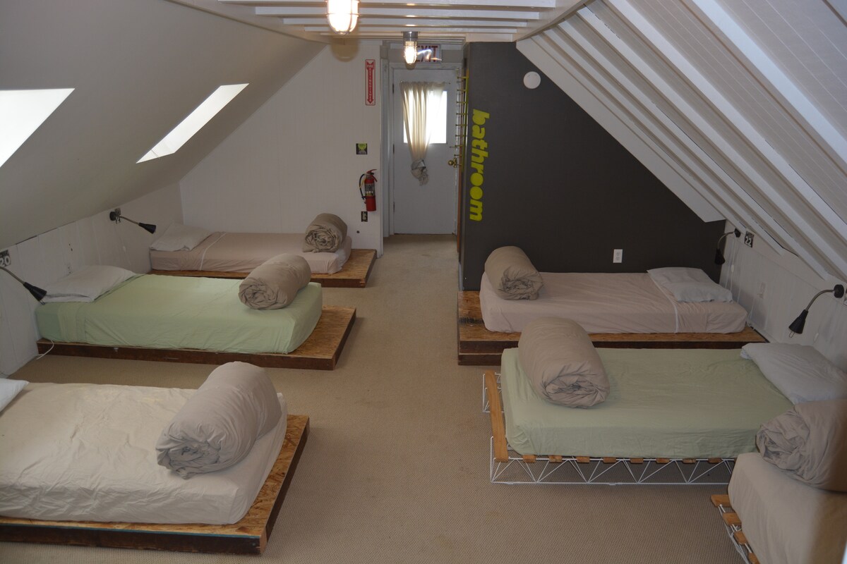 A spacious dormitory room is equipped with eight single beds, each adorned with simple bedding in various hues. Soft lighting is provided by wall-mounted lamps next to the beds. A bathroom door is visible at the far end, and sloped ceilings enhance the room's airy feel.