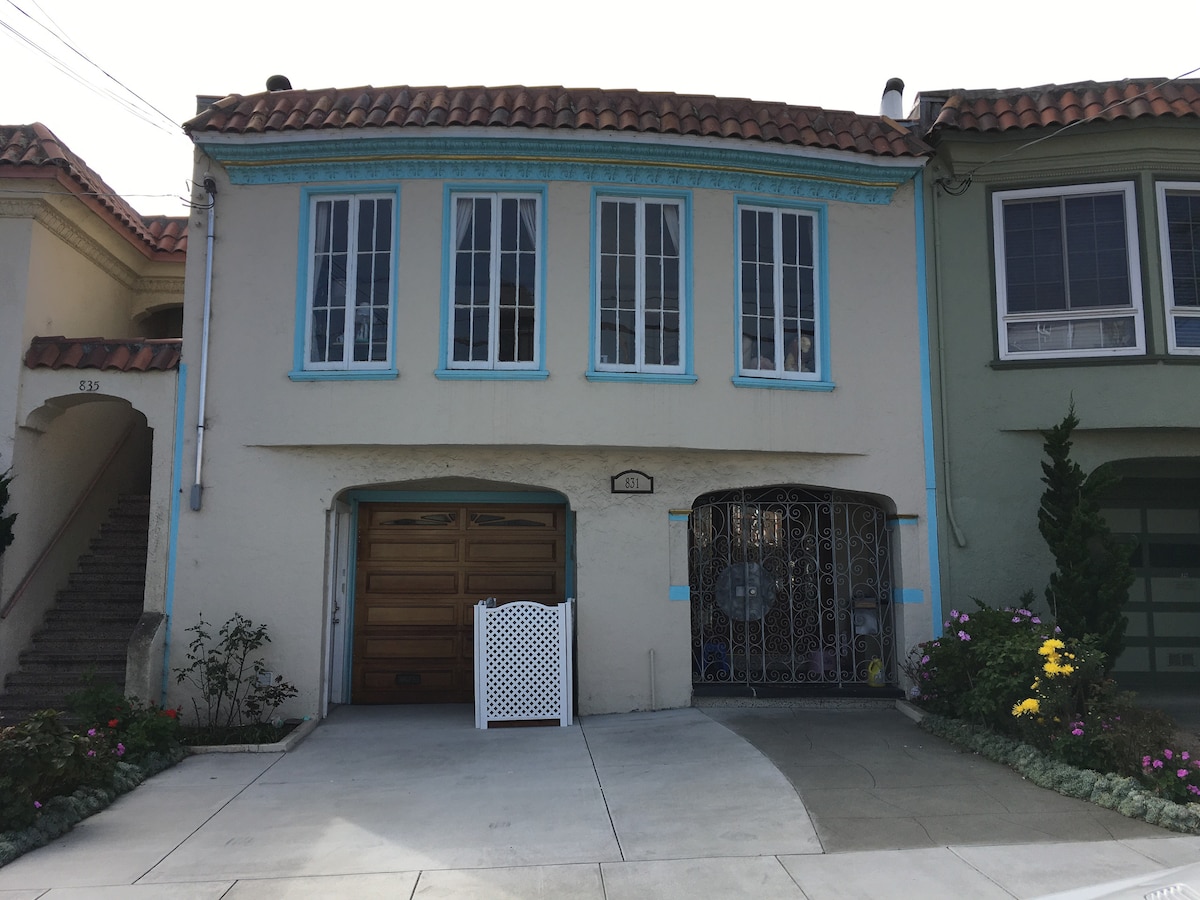 A two-story residential building with a textured façade painted in light beige, framed by a blue decorative trim. Large windows with multiple panes allow natural light to enter. The driveway features a garage door and is bordered by landscaped flower beds, adding color and vibrancy.