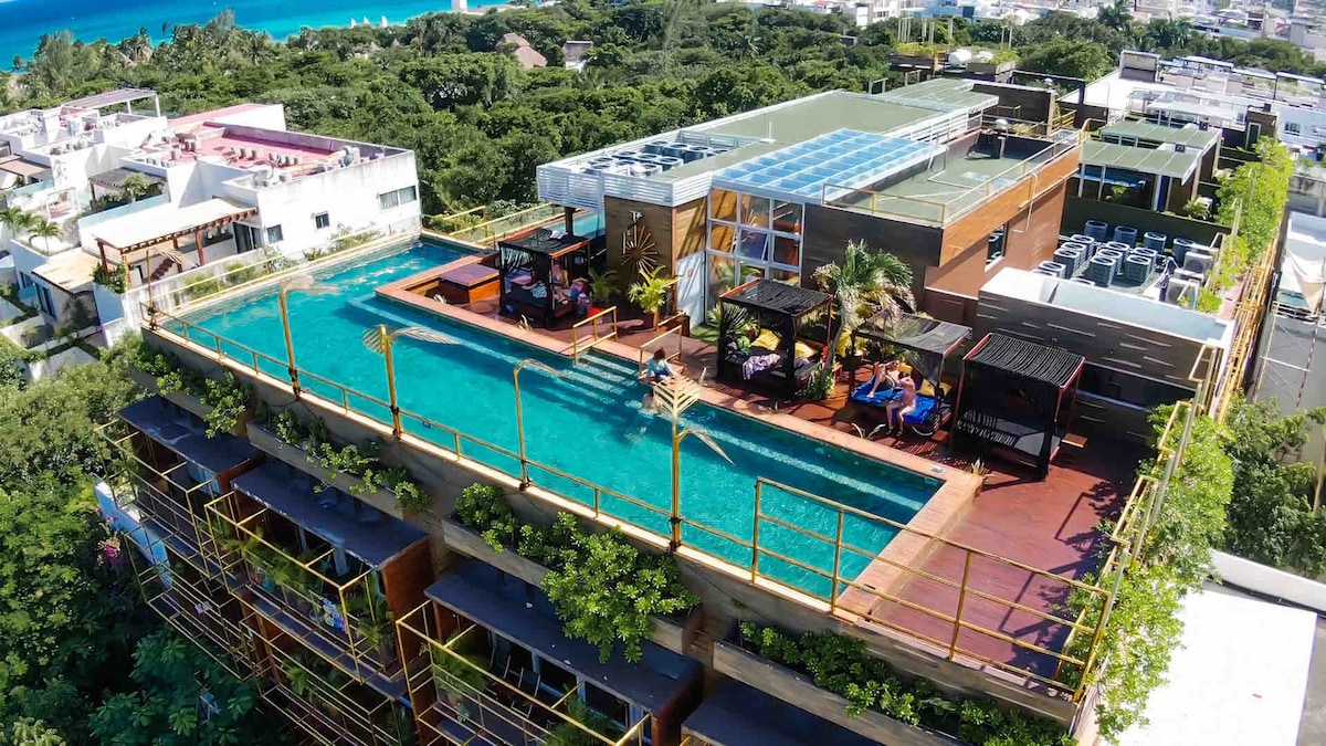 An aerial view of the rooftop area features two swimming pools surrounded by lounge areas with shaded seating. Lush greenery adorns the edges of the building, enhancing the connection with nature. The ocean can be glimpsed in the background, adding to the serene atmosphere.