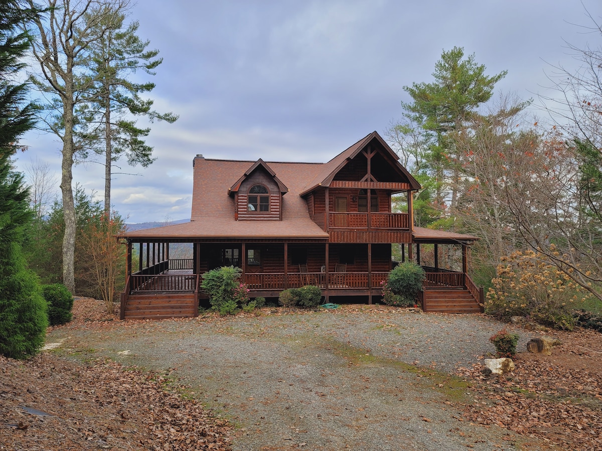 A three-story cabin is nestled among trees, featuring a broad front porch with multiple entrance points. The exterior is finished in warm wood tones, complemented by a peaked roof and decorative gables. Surrounding foliage adds a natural touch to the landscape.